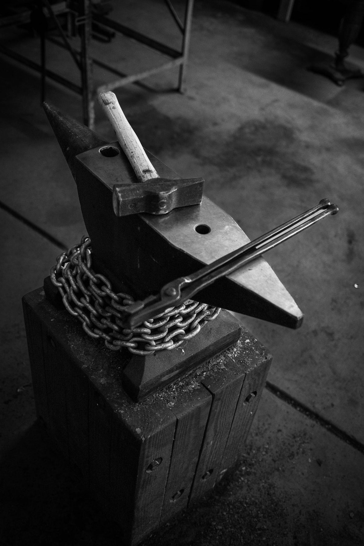 A black and white photograph of a heavy forge anvil with a chain wrapped around its base, holding a piece of metal, with a hammer resting on top, set in a dark workshop environment.